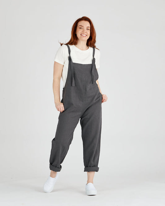 Dark grey linen overall, front view on female model with hands in pockets. 